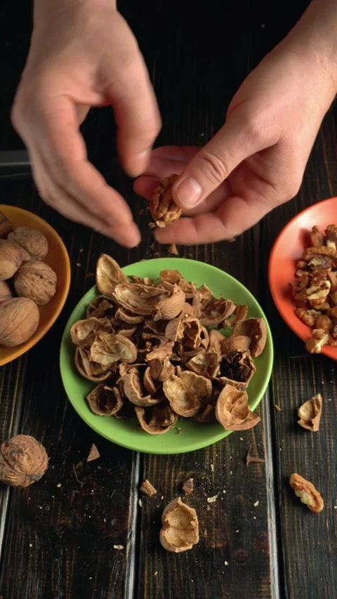 Hands cracking open walnut shells over a green bowl filled with empty shells, Stock Footage 335597534