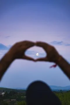 Hands create a heart shape framing the moon during a serene twilight sky over Stock Photos