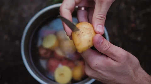 Hands cuts potatoe in half, two slices drops in pot of water, field kitchen Stock Footage 137591347