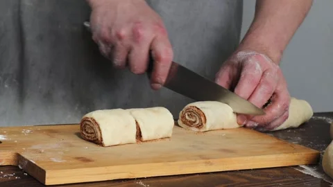 Hands cutting cinnamon roll dough log into portions on wooden board during Stock Footage 310337439