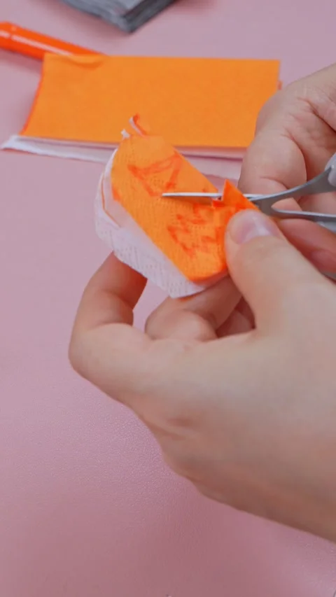 Hands cutting orange sticky note paper with small scissors on a pink table Видео 346960050