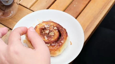 Hands delicately pulling apart a cinnamon roll on a cozy wooden table Stock Footage 317461572