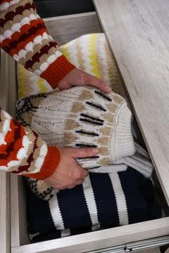 Hands folding cozy knitted sweaters inside wooden dresser drawer closeup Stock Photos