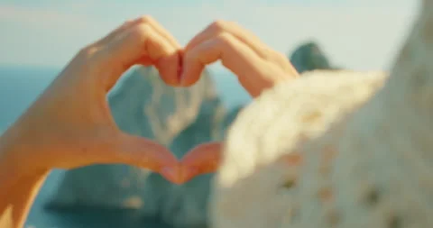 Hands forming heart shape framing distant Faraglioni sea cliffs over calm blue Stock Footage 341292691