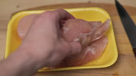 Hands gently handle raw chicken breast on a yellow tray in a kitchen setting. Видео 338298245
