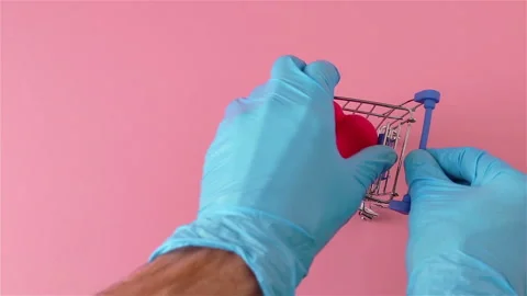 Hands in gloves take red heart from small shopping cart and put on pink surface Stock Footage 147537474