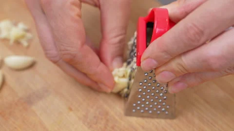 Hands grating fresh garlic on a mini red grater over a wooden board Видео 346799715