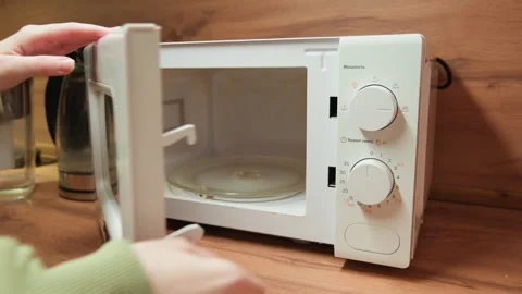 Hands in green sleeves open white microwave door and turn lower dial on wooden Stock Footage 343536108