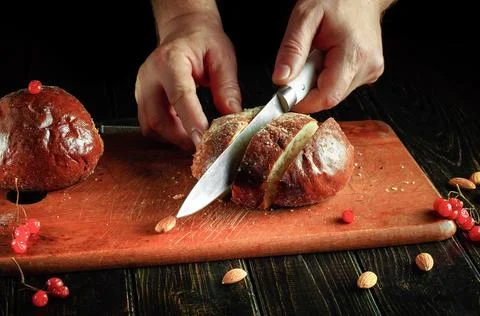 Hands hold a knife to slice bread on a wooden cutting board. Red berries an.. Stock Photos