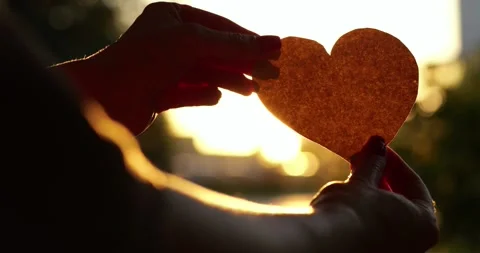 Hands hold paper heart at sunset. Golden light creates romantic silhouette Stock Footage 346690175