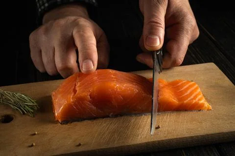Hands hold scissors as they cut a salmon fillet on a wooden board. The settin Stock Photos