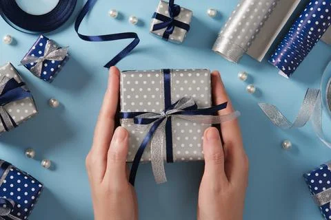 Hands hold a silver-colored gift box. Around it are gifts and ribbons in blue Stock Photos