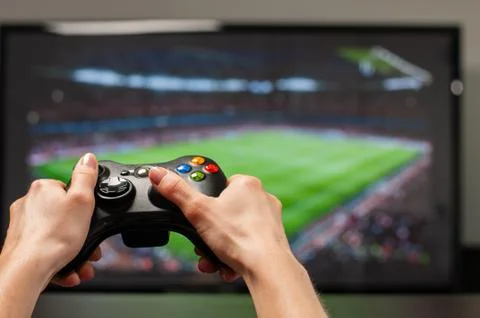 Hands holding the game controller while man playing football game on TV Stock Photos