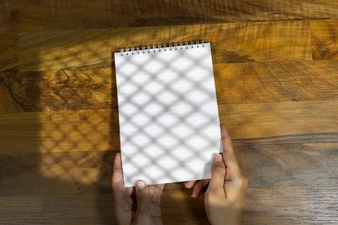 Hands Holding Small Spiral Notepad Over Wooden Table with Dappled Shadow Pattern Stock Photos