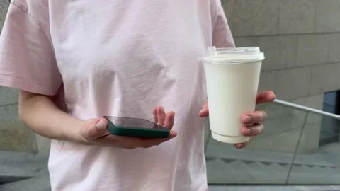 Hands holding smartphone and white paper coffee cup, person in pink t-shirt Видео 340149141