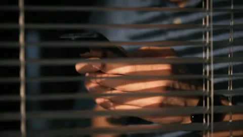 Hands holding a smartphone behind window blinds in a low-light environment Stock Footage 323806696