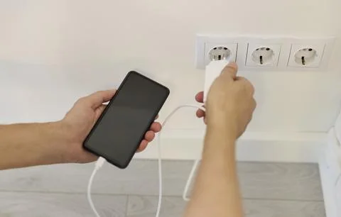 Hands holding smartphone while inserting charger into wall socket with white Stock Photos