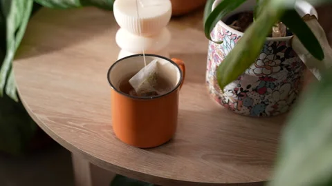 Hands Infusing a Tea Bag in an Orange Mug on a Nightstand Surrounded by Plants Stock Footage 302820696