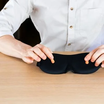 Hands inserting orange earplugs into black sleep mask on table. Stock Photos