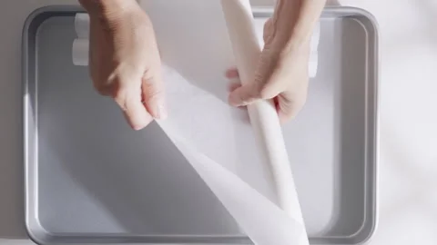 Hands lay a large sheet of parchment paper over an empty metal baking tray.  Stock Footage 321849664