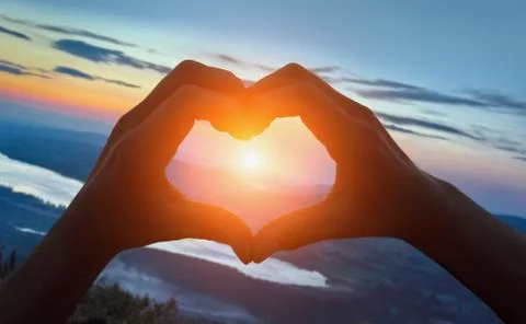 Hands making heart shape on the beach against sunrise over mountains Stock Photos