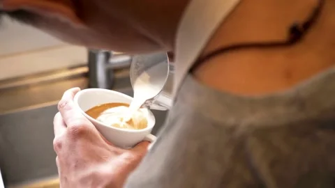 Hands making latte art at a cafe. Making Latte Art by pouring milk into coffee. 動画素材 334648451