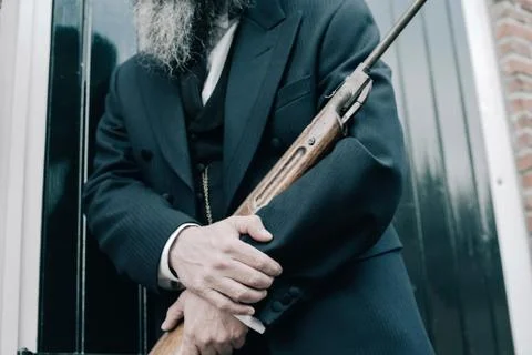 Hands of man holding rifle while leaning against window shutters. Stock Photos