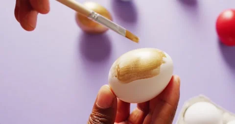 Hands painting white eggs using brush with gold paint on purple background 스톡 동영상 331246026