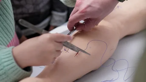 Hands performing stitching practice on flesh toned mannequin leg with forceps Stock Footage 344873973