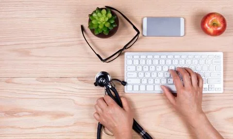 Hands picking up stethoscope while using computer keyboard on wooden desktop Stock Photos