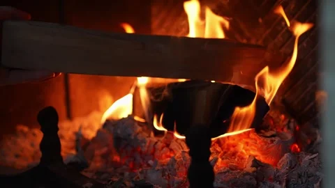 Hands place a log onto glowing embers in a cozy fireplace as flames rise and Stock Footage 328132406