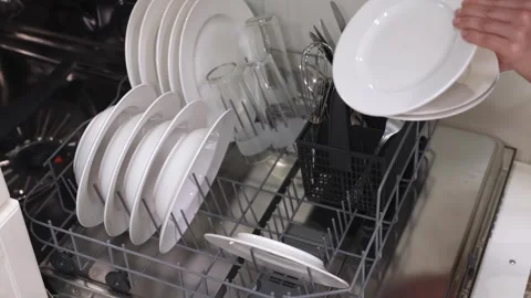Hands placing clean white plates into a dishwasher rack alongside glasses and Stock Footage 324309310