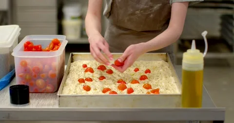 Hands placing halved cherry tomatoes into dimpled focaccia dough in commercial Stock Footage 344595523