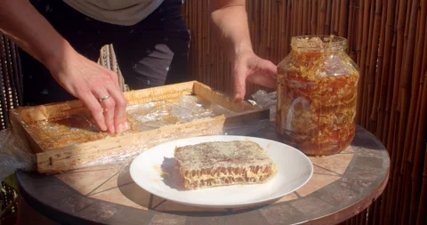 Hands pressing honeycomb comb and tasting honey from jar on sunlit patio table Stock Footage 344884638