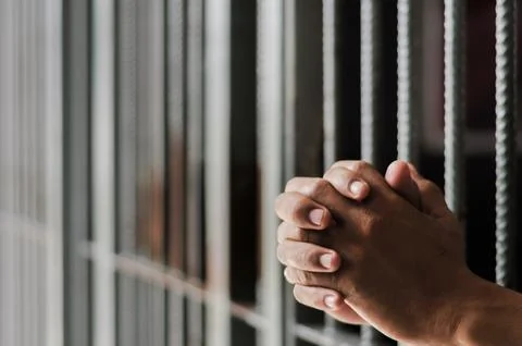 Hands of a prisoner behind prison bars on black background Stock Photos