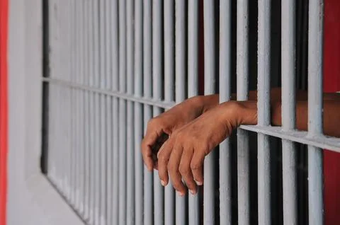 Hands of a prisoner behind prison bars on black background Stock Photos