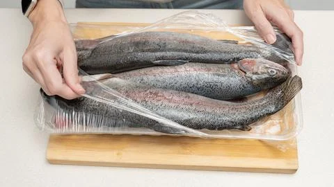 Hands removing plastic wrap from fresh rainbow trout fish on a wooden cutting Stock Photos