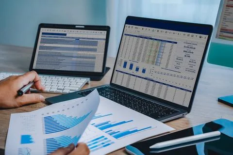 Hands reviewing financial chart document with laptop displaying spreadsheet.. Stock Photos