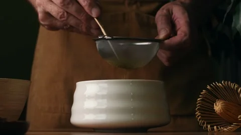 Hands sift roasted hojicha powder through fine mesh sieve into ceramic bowl Video stock 340874439