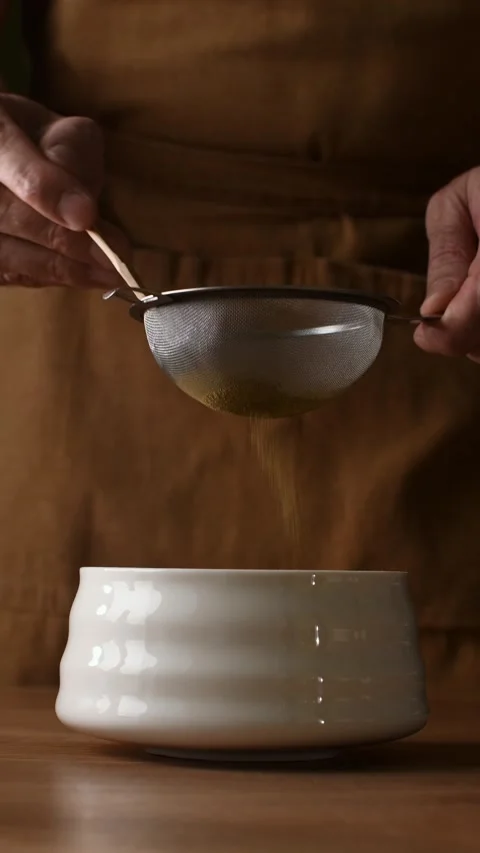 Hands sift roasted hojicha powder through fine mesh sieve into ceramic bowl Video stock 340874770