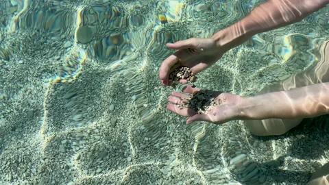 Hands sifting through small pebbles in crystal clear turquoise water on a sunny Stock Footage 315293744