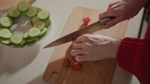 Hands slicing cherry tomatoes beside cucumber rounds on wooden board in home Stock Footage 346642728