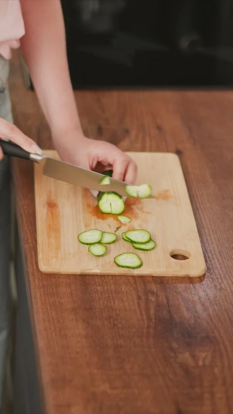 Hands slicing fresh cucumber into thin rounds on wooden cutting board with knife Stock Footage 346776381