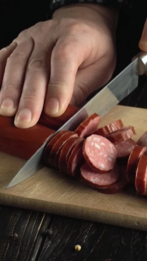 Hands slicing sausage into even rounds on a wooden cutting board with a sharp Video stock 338306667