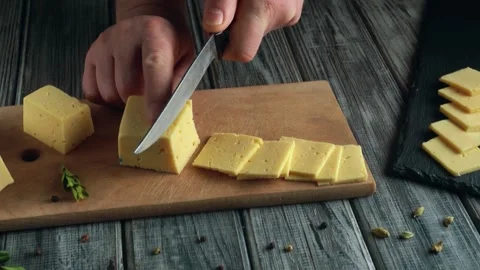 Hands slicing yellow cheese into cubes and thin slices on a wooden cutting board Stock Footage 337374520