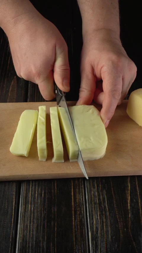 Hands slicing yellow cheese into even pieces on a wooden cutting board, Vidéo 328090598