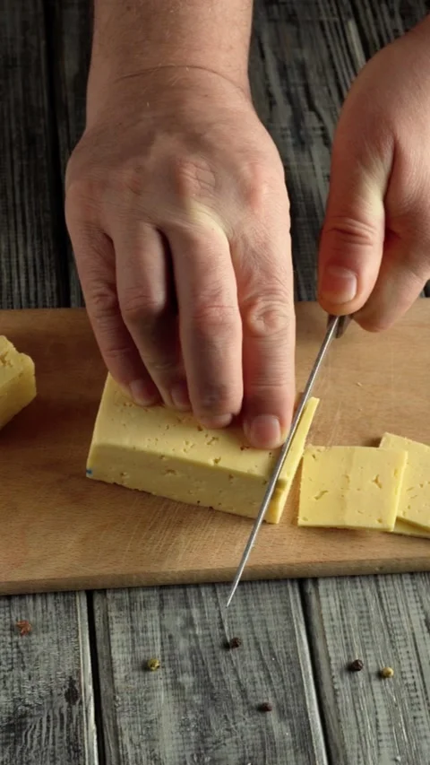 Hands slicing yellow cheese into even pieces on a wooden cutting board with a Stock Footage 337696650