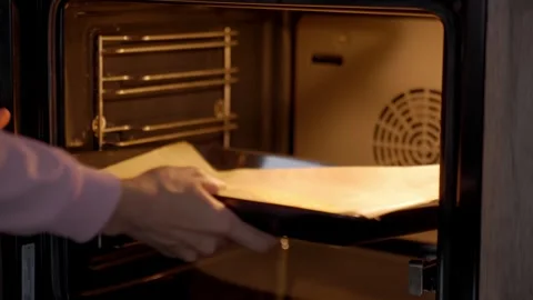 Hands slide parchment lined baking tray into warm oven during home baking Stock Footage 340854415