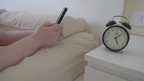 Hands with a smartphone in bed next to a retro alarm clock. Stock Footage 320573450