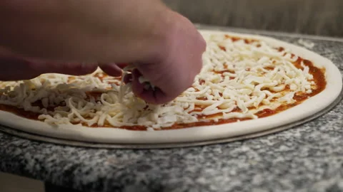 Hands sprinkle shredded mozzarella cheese over red sauce on raw pizza, Vídeo Stock 217778807